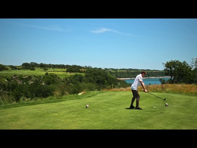 Man swings a golf club on a green tee box overlooking a scenic blue lake.